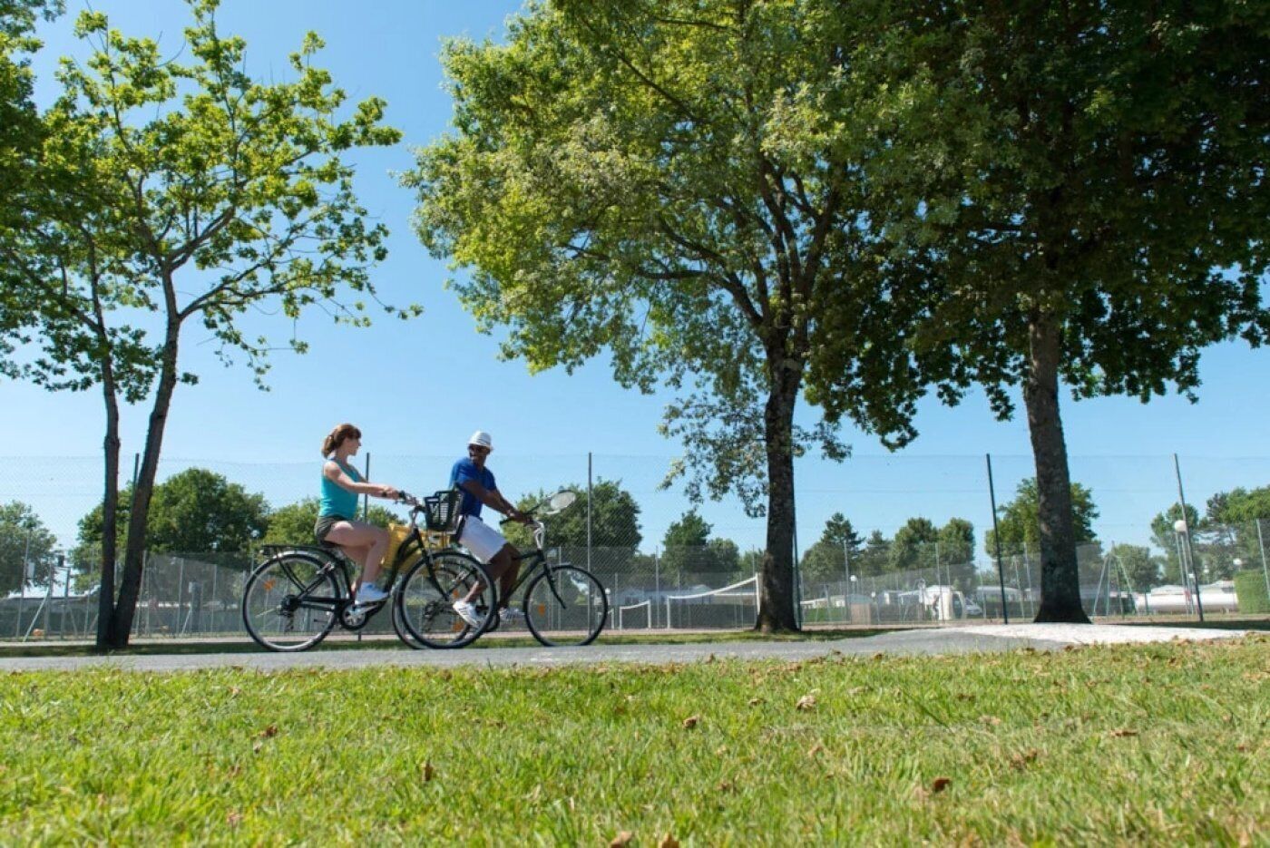 Two people riding bicycles on a path with trees and a fence in the background.