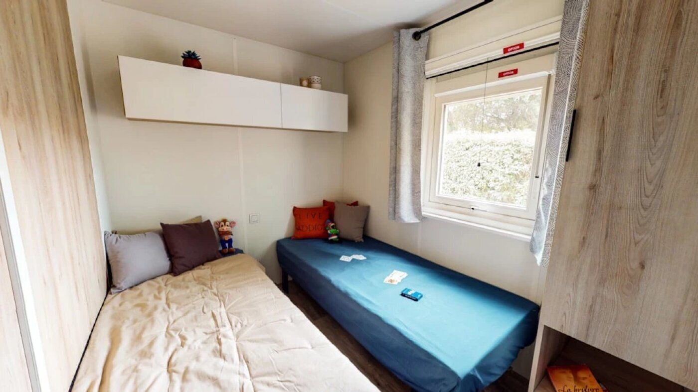 Una habitación pequeña con cama, una mesita, una ventana con cortinas y un estante sobre la cama.