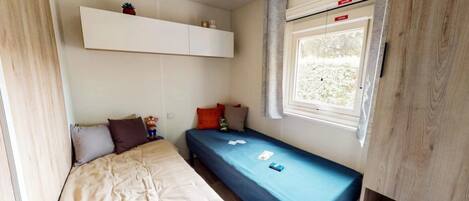Una habitación pequeña con cama, una mesita, una ventana con cortinas y un estante sobre la cama.