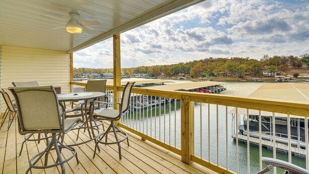 A wooden deck with a table and chairs, overlooking a river with boats and a distant shoreline. - Lakefront Osage Beach Condo w/ Water-View Balcony! (Osage Beach)