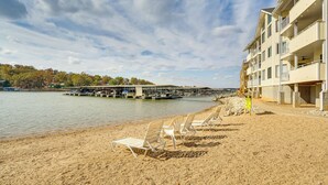 A beach with white lounge chairs and a building with balconies. - Lakefront Osage Beach Condo w/ Water-View Balcony! (Osage Beach)