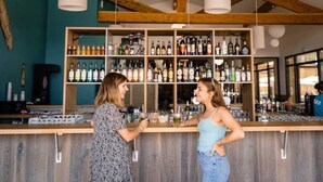 A bar area with wooden shelves stocked with various bottles and glasses, a counter with a person serving, and a rustic ceiling with exposed beams. - ZEcamping 3* toboggans club enfant locatif clim 28m2 6 pers (Saint-Julien-en-Born)