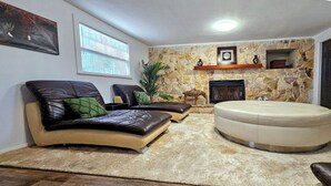 A living room with a stone wall, a fireplace, a large round ottoman, and a leather sofa. - 2 Mi to Cocoa Riverfront Park: Home w/ Patio! (Cocoa)