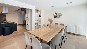 Ett modernt matområde med ett träbord och grå stolar, ett köksområde med en inbyggd köksö och en vit vägg med en dekorativ spegel.