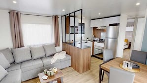 A modern living room with a sofa, a coffee table, and a dining area with a table and chairs. There is a kitchen area with a refrigerator and a stove. - ZEcamping 4 étoiles animé 1km plage toboggans locatif TV LV clim 36m2 (Les Mathes)