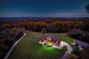 A large, two-story house with a well-maintained lawn and driveway, surrounded by trees and a clear night sky. - Luxe Bourbon Trail Cabin– Hot Tub/Pickleball/Fire Pit/Pool/Game Room (Lawrenceburg)