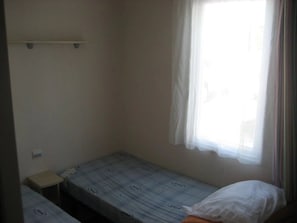 Une petite chambre avec un lit simple, une fenêtre, une table de chevet et une étagère au-dessus du lit.