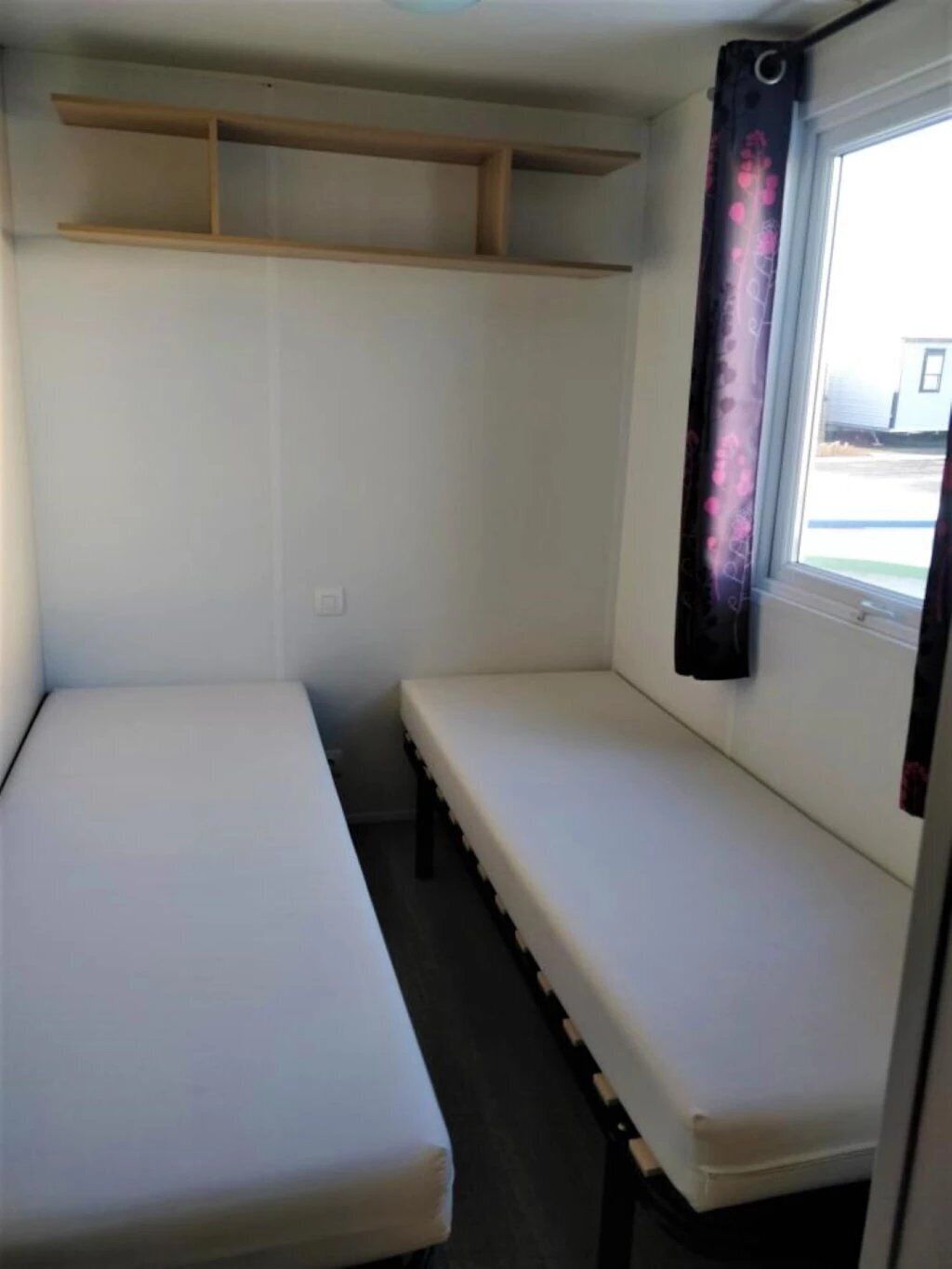 A small, single-bed room with a window, a curtain, and a wooden shelf above the bed.