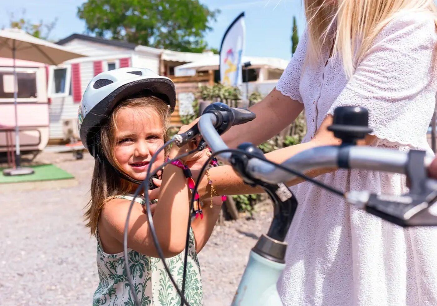 A person assisting a child with a bicycle in an outdoor setting with caravans in the background.