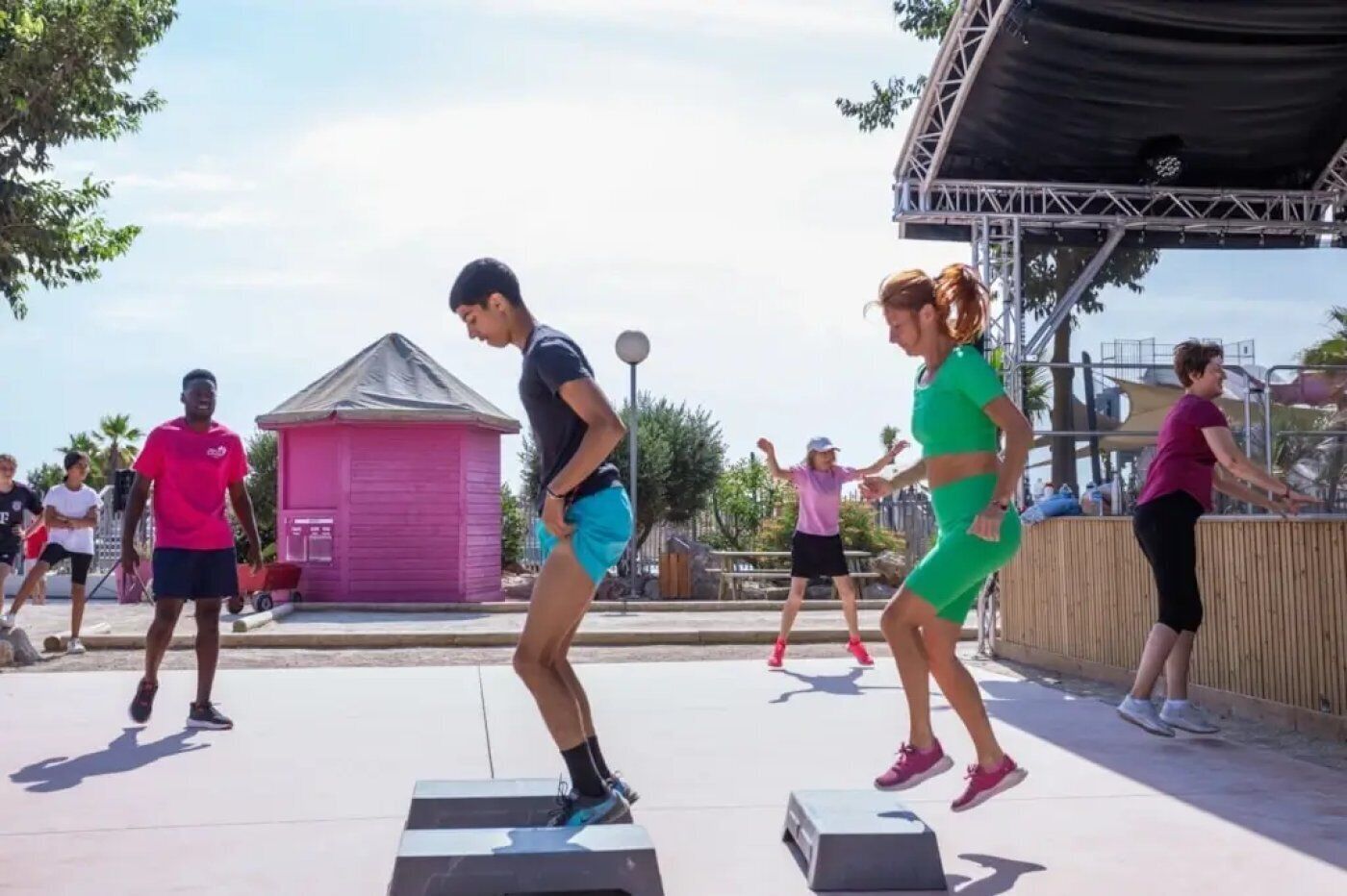 A group of people exercising outdoors with a stage and a small building in the background.