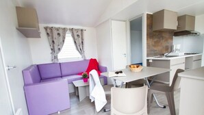 A compact living space with a purple sofa, a small dining table, a kitchen area, and a window with curtains. - ZEcamping 4 étoiles animé parc aquatique locatif TV 23m2 4 pers (Valras-Plage)