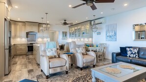 A spacious living room with a large glass coffee table, two beige armchairs, a blue sofa, and a chandelier.