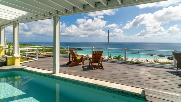 A pool area with a view of the ocean and beach, wooden deck, and two Adirondack chairs.