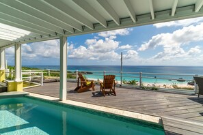 Zona de piscina con vista al océano y a la playa, terraza de madera y dos sillas Adirondack.