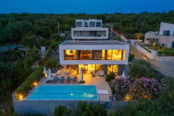 Una casa moderna de dos pisos con piscina, mobiliario exterior y vistas a la vegetación circundante.