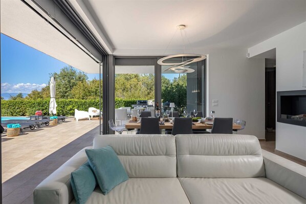 A modern living room with a large sofa, a dining area with a table and chairs, and a view of a pool and greenery outside.