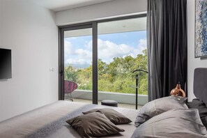 Een moderne slaapkamer met een groot bed, een flatscreen televisie en een balkon met uitzicht op groen en de lucht.