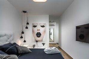 A modern bedroom with a tufted headboard, a bed with dark bedding, a large wall-mounted artwork, and a TV on the wall.