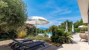 A poolside area with lounge chairs and umbrellas, surrounded by greenery and a view of a pool and mountains.