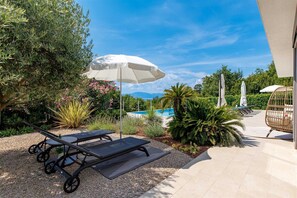 Zona junto a la piscina con sillones de descanso y sombrillas, rodeada de vegetación y vistas a una piscina y montañas.