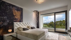 A modern bedroom with a large bed, a view of greenery through a sliding glass door, and a decorative wall piece.