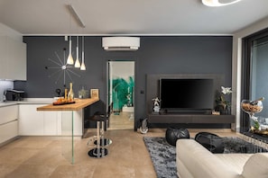 A modern living room with a large flat-screen TV, a dining area with a wooden table, and a kitchen with a white cabinet.