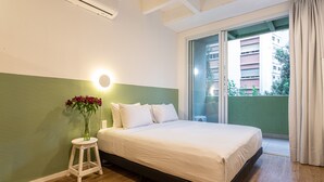 Ein Schlafzimmer mit einem Bett, einem Nachttisch mit Blumenvase, einer Wandleuchte und Blick auf Gebäude durch ein Fenster.
