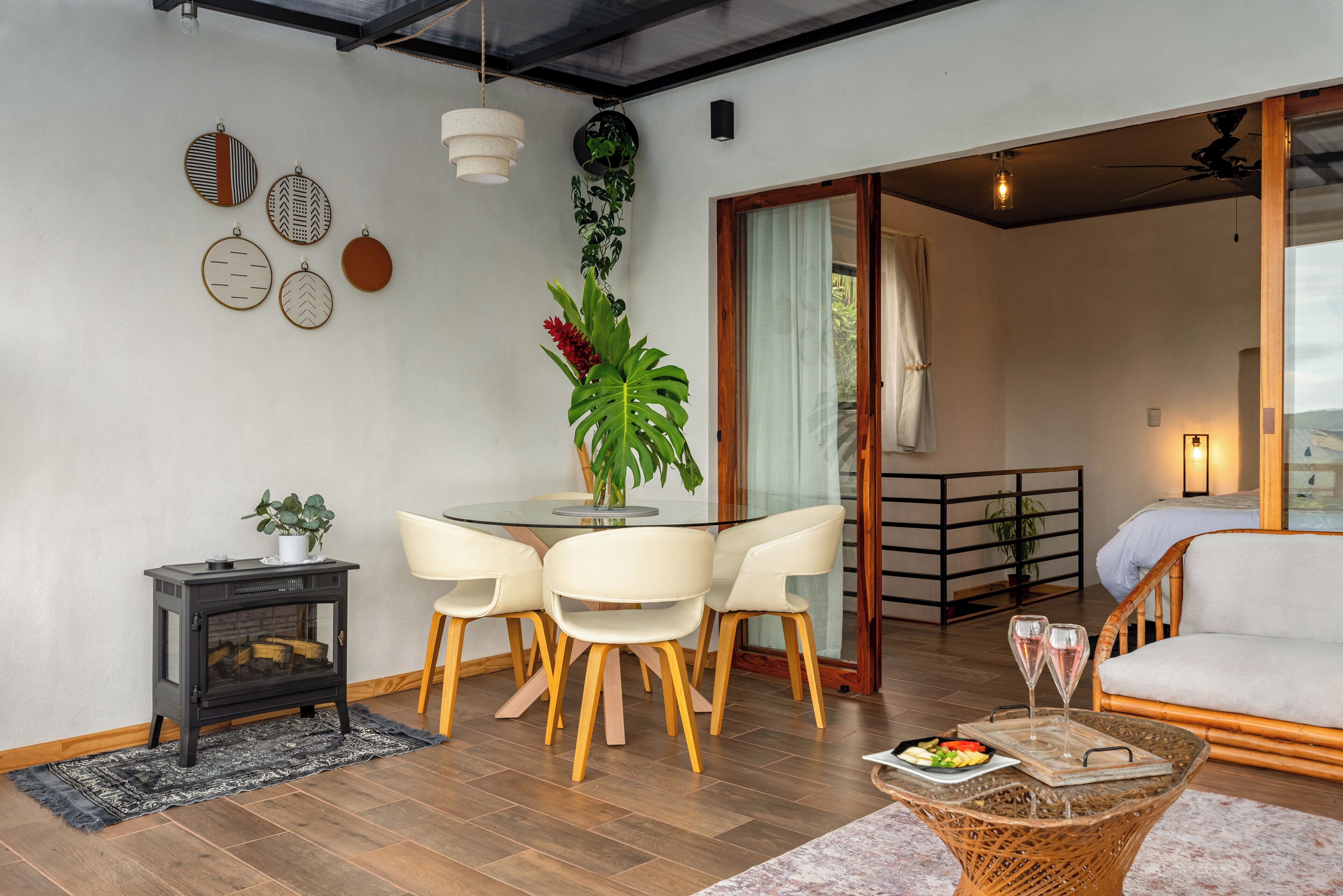 A cozy living room with a glass-top dining table, white chairs, a black wood-burning stove, and a wicker coffee table with a tray of food and glasses.