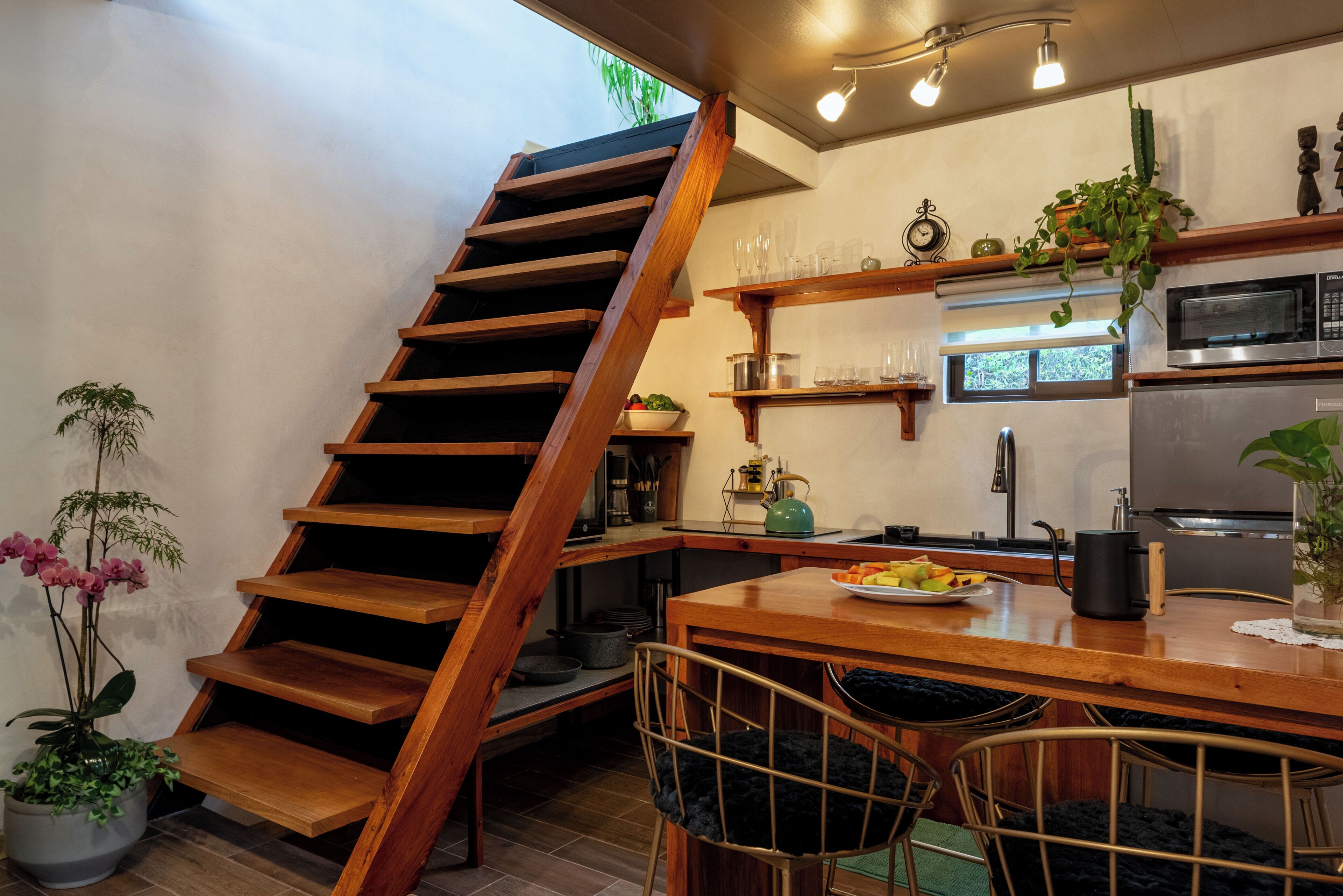 A kitchen with a wooden staircase, a dining area with chairs, and a kitchen counter with a plate of fruit.