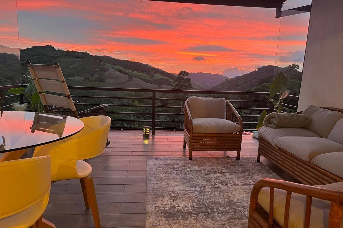 A balcony with a glass table and yellow chairs, a wicker sofa, and a view of a sunset over hills.