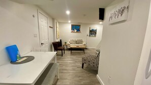 Un salon moderne avec un canapé blanc, un fauteuil à motifs et une table basse. On y trouve une table à manger avec un banc et un dossier bleu posé dessus. La pièce est dotée d’un plancher en bois et de murs blancs.