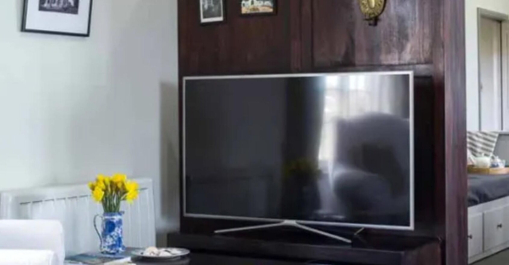 Un salon avec un téléviseur à écran plat posé sur un meuble en bois, un vase rempli de fleurs jaunes et une chaise blanche.
