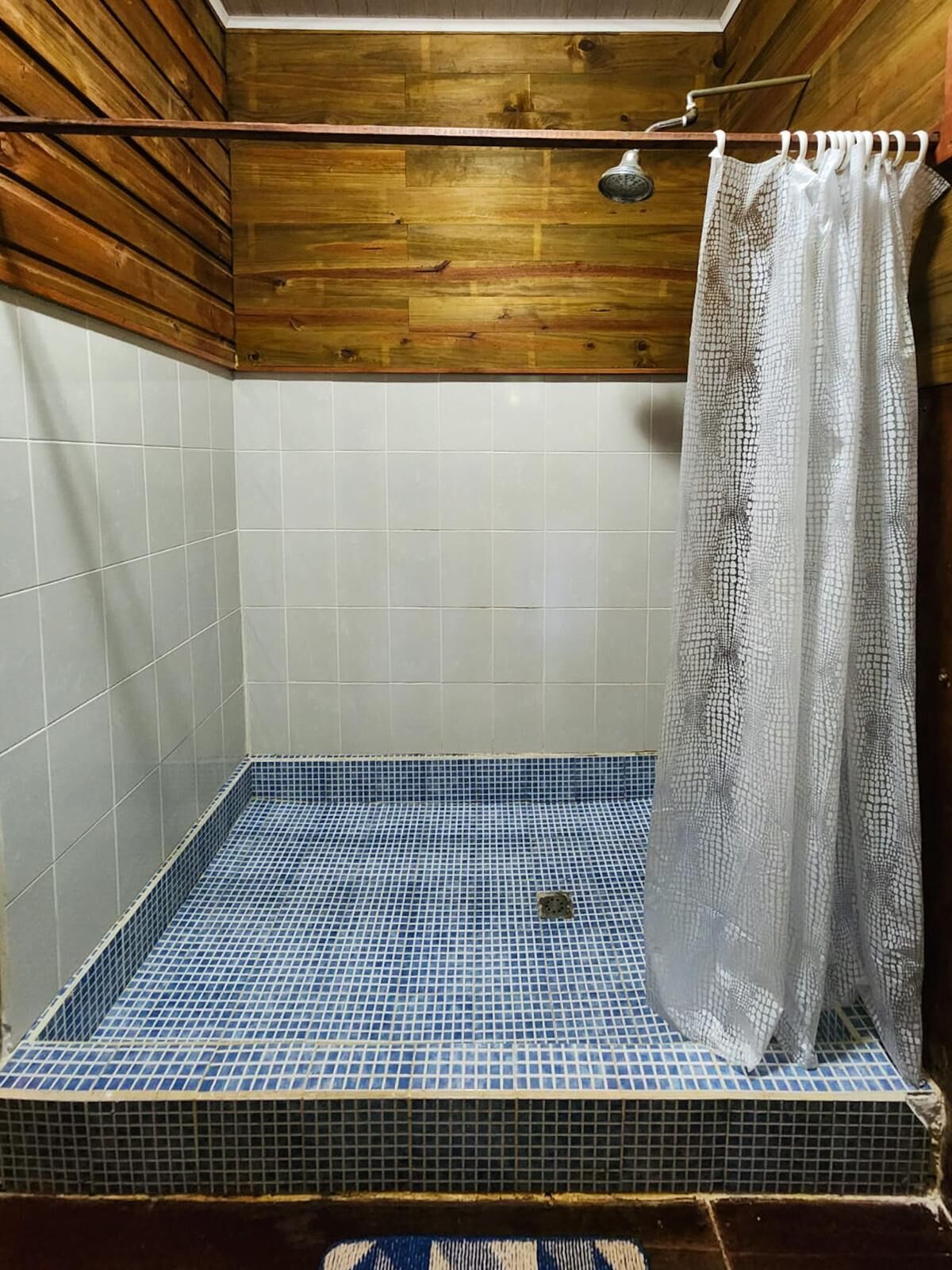 Een douche met een blauw betegelde vloer, houten plafond en een wit douchegordijn.