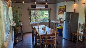 A rustic dining area with a wooden table and chairs, a window with a view of greenery, a kitchen area with a refrigerator, and hanging lights.