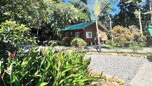 A single-story wooden house with a green roof, surrounded by lush greenery and trees.