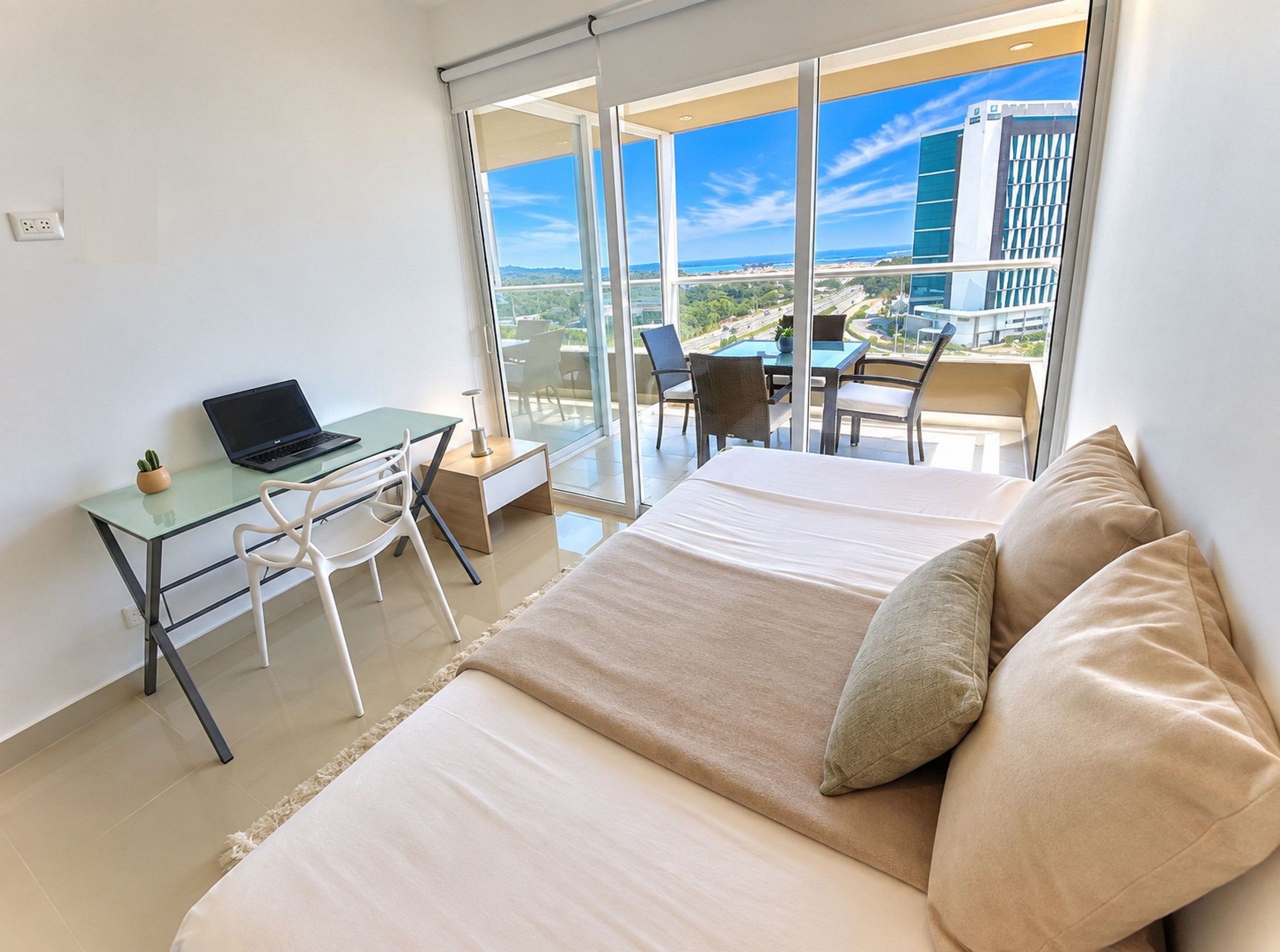 A modern hotel room with a bed, a desk with a laptop, and a balcony with a table and chairs.