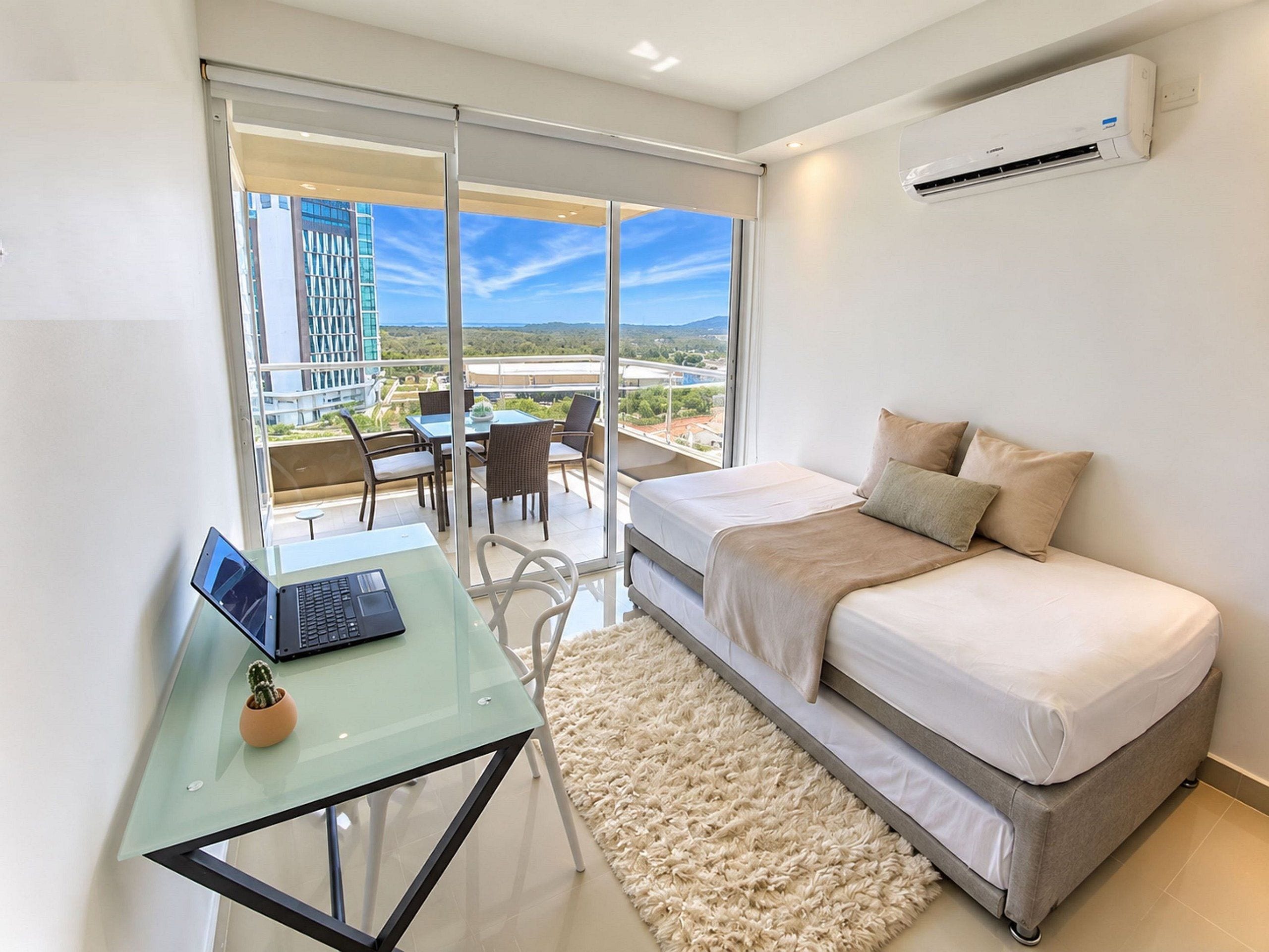 Un dormitorio moderno con una cama, una computadora portátil sobre una mesa de vidrio y vistas a un balcón con mobiliario exterior.