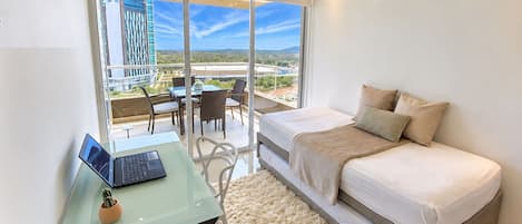 A modern bedroom with a bed, a laptop on a glass table, and a view of a balcony with outdoor furniture.