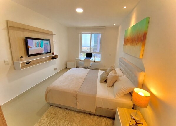 A modern bedroom with a bed, a TV mounted on the wall, a desk with a laptop, and a window with a view.