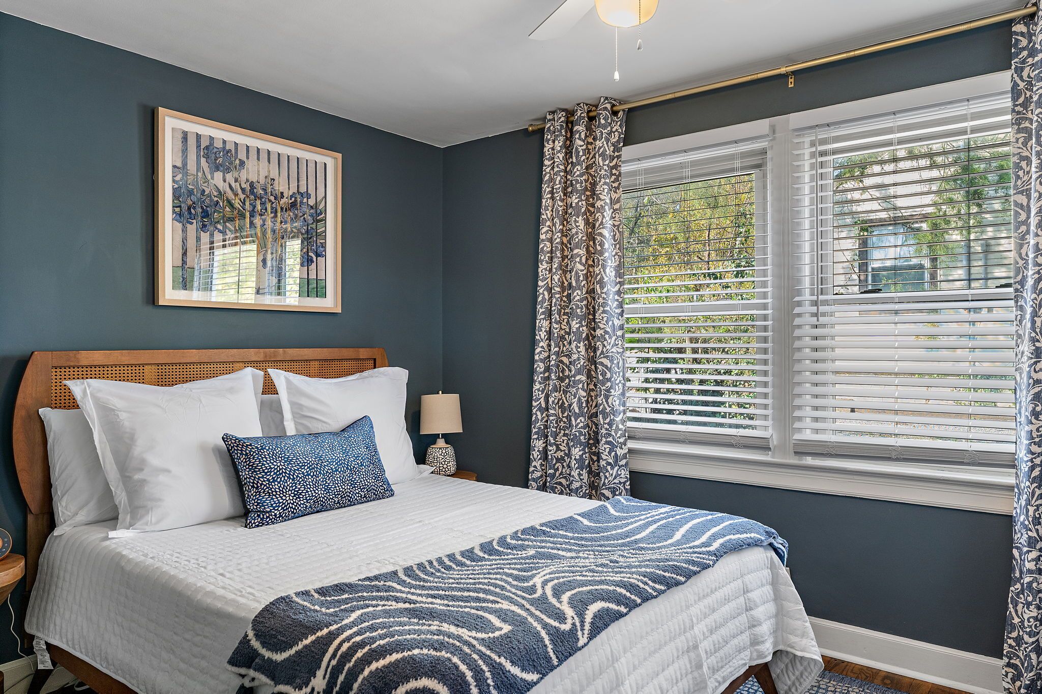 A bedroom with a bed, a nightstand, a lamp, a window with blinds, and a framed picture on the wall.