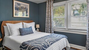 A bedroom with a bed, a nightstand, a lamp, a window with blinds, and a framed picture on the wall.