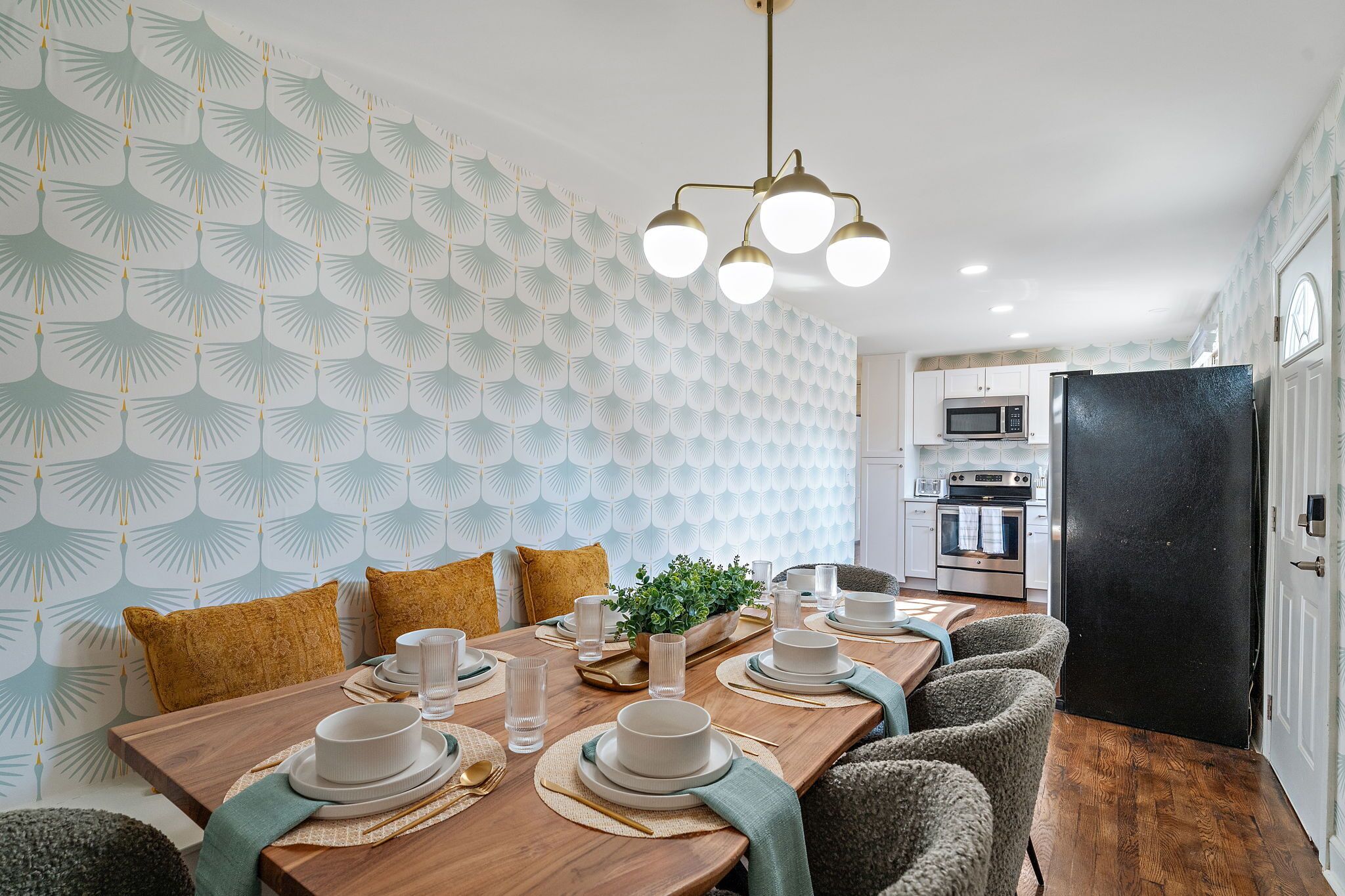A dining area with a wooden table set for four, surrounded by chairs. A modern chandelier hangs above, and the room features patterned wallpaper and a black refrigerator.
