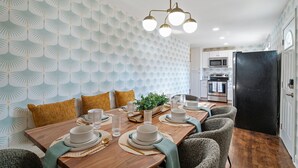 A dining area with a wooden table set for four, surrounded by chairs. A modern chandelier hangs above, and the room features patterned wallpaper and a black refrigerator.