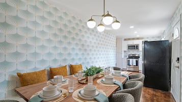 A dining area with a wooden table set for four, surrounded by chairs. A modern chandelier hangs above, and the room features patterned wallpaper and a black refrigerator.