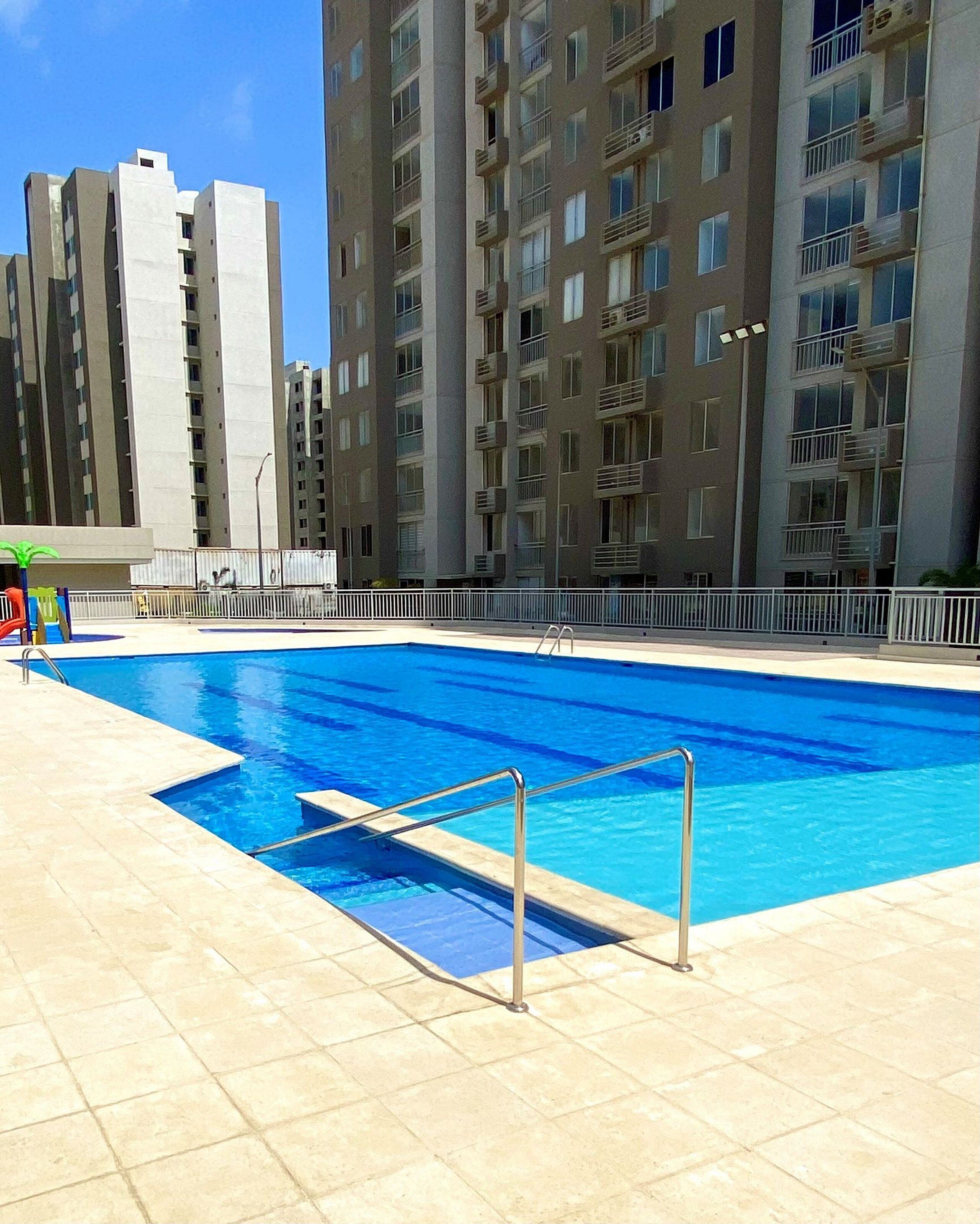 A swimming pool with a metal ladder, surrounded by a tiled deck, and adjacent to high-rise residential buildings.