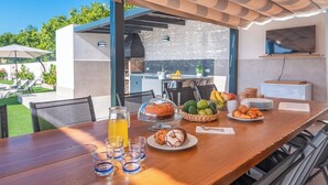 Ein Holzespeisetisch ist reichhaltig mit Speisen und Getränken gedeckt, ein Fernseher ist an der Wand befestigt, und es gibt Sitzgelegenheiten im Freien mit Sonnenschirmen.