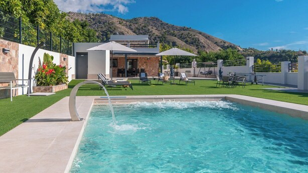 Ein Schwimmbad mit Wasserspiel, umgeben von einer Terrasse mit Stühlen und Sonnenschirmen, mit Hügeln im Hintergrund.
