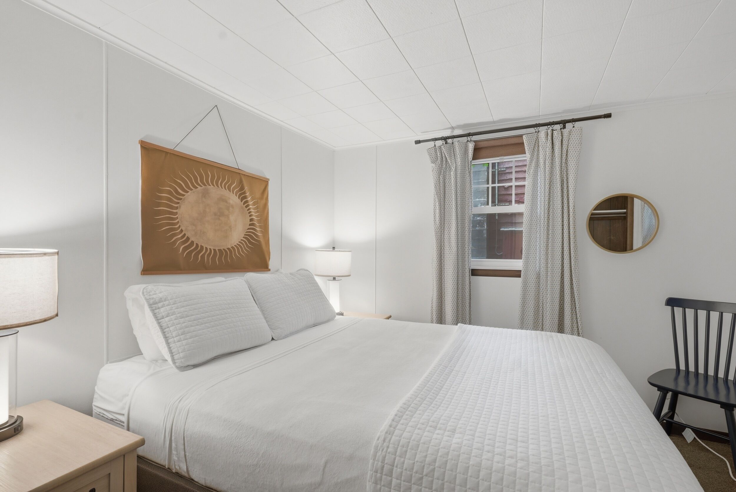 Ein Schlafzimmer mit einem Bett, einem Nachttisch, einer Lampe, einem wandmontierten Spiegel und einem Fenster mit Vorhängen.