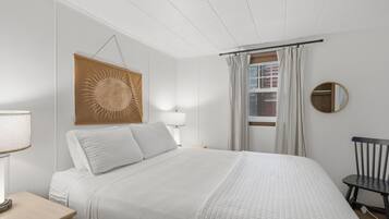 Ein Schlafzimmer mit einem Bett, einem Nachttisch, einer Lampe, einem wandmontierten Spiegel und einem Fenster mit Vorhängen.