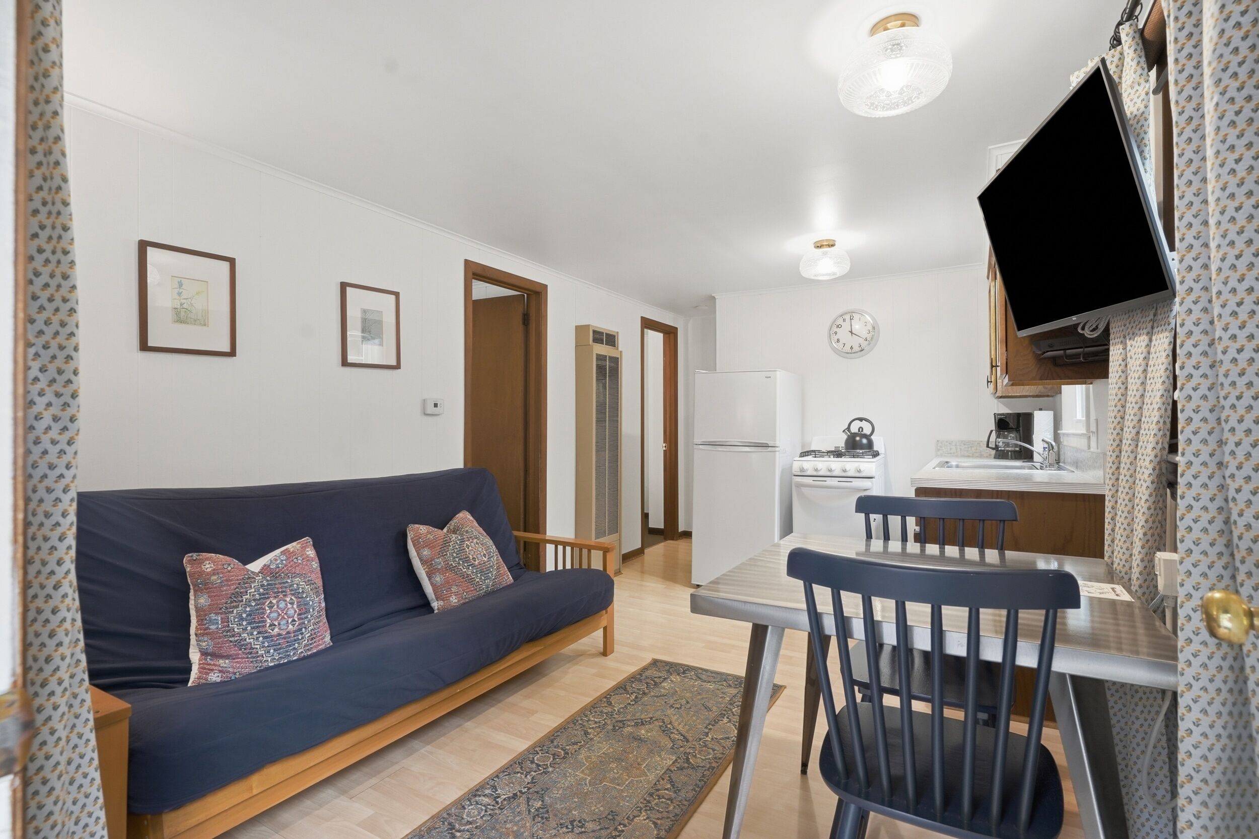A compact living space with a sofa, dining table, chairs, a TV, kitchen area, and a patterned rug.